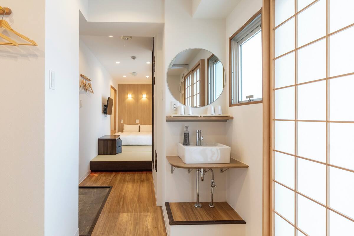A hotel hallway with a bed, a bathroom with a sink and mirror, and wooden flooring.