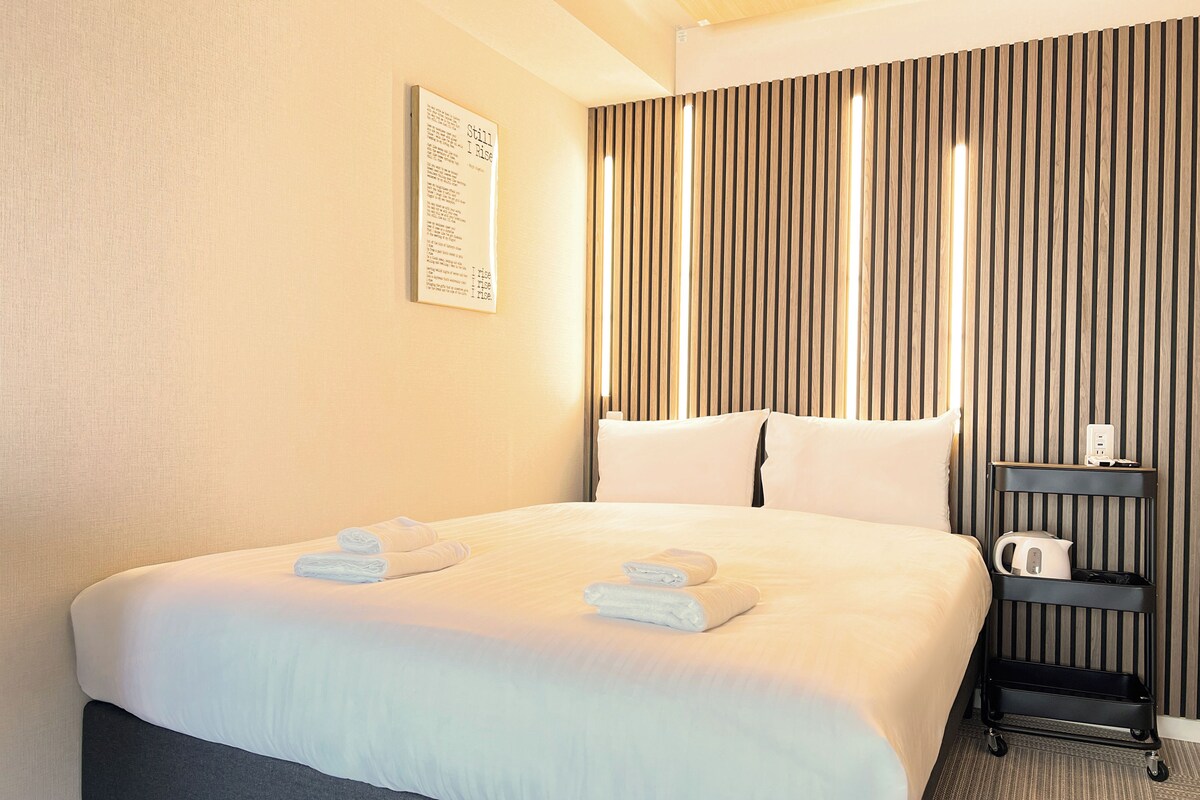 A hotel room with a large bed, a bedside table with a kettle, and a wall-mounted sign.