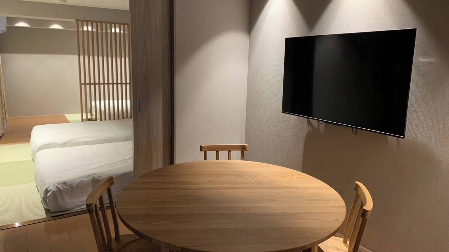 A room with a bed, a round wooden table, two chairs, a TV mounted on the wall, and a window with curtains.