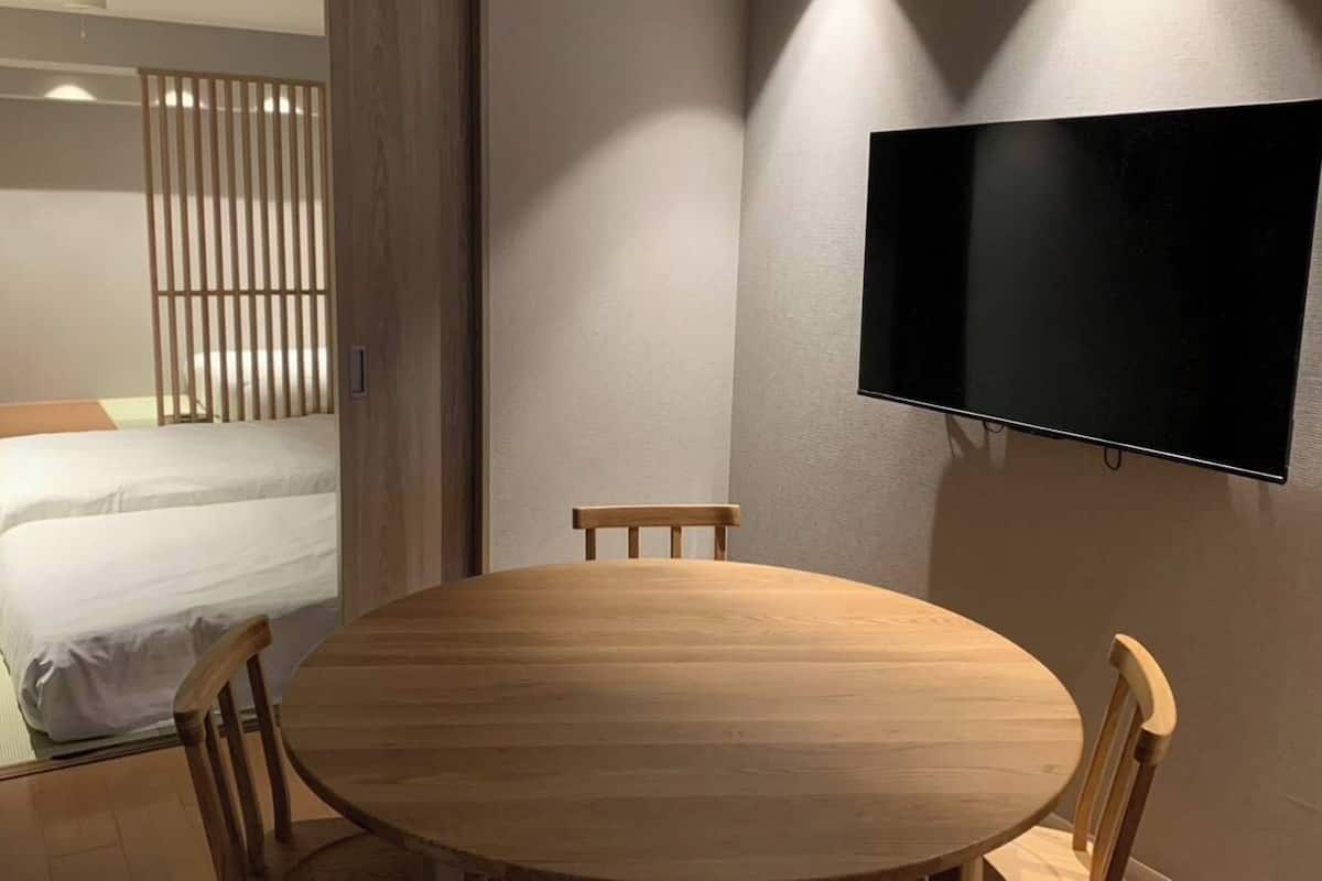A room with a bed, a round wooden table, two chairs, a TV mounted on the wall, and a window with curtains.