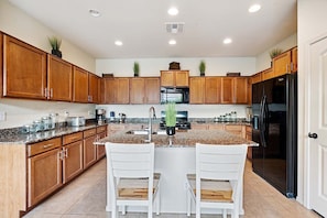 A kitchen with wooden cabinets, granite countertops, a black refrigerator, and two white bar stools. - Beautifully Furnished Home in Power Ranch ~ Steps away from Community Pool & Spa (Gilbert)