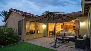 A patio area with a dining table and chairs, a large umbrella, and a view into the house. - Beautifully Furnished Home in Power Ranch ~ Steps away from Community Pool & Spa (Gilbert)