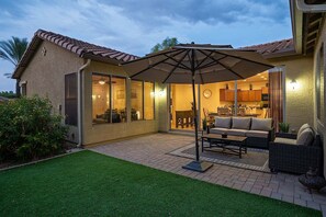 A patio area with a dining table and chairs, a large umbrella, and a view into the house. - Beautifully Furnished Home in Power Ranch ~ Steps away from Community Pool & Spa (Gilbert)