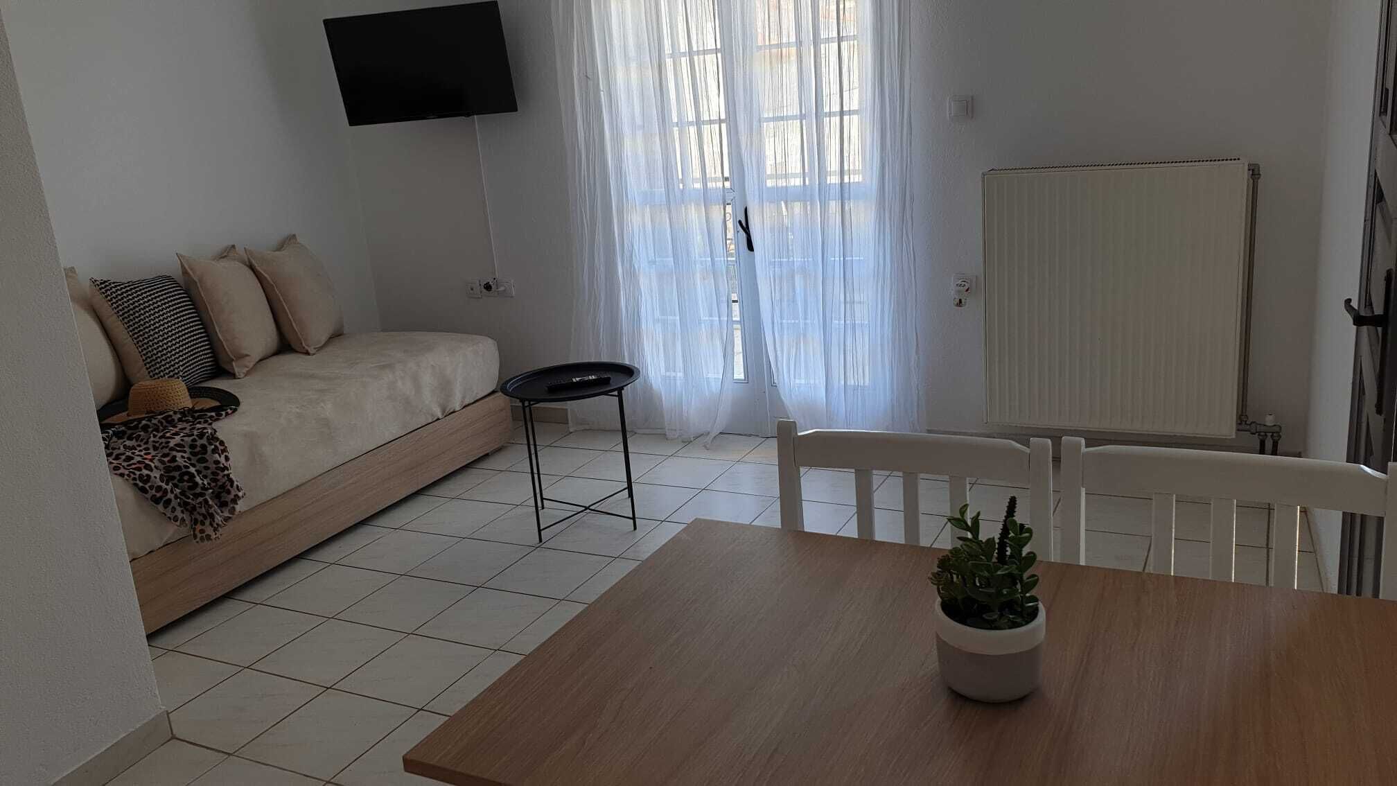 A minimalist living room with a sofa, a small table, a TV mounted on the wall, and a window with sheer curtains.