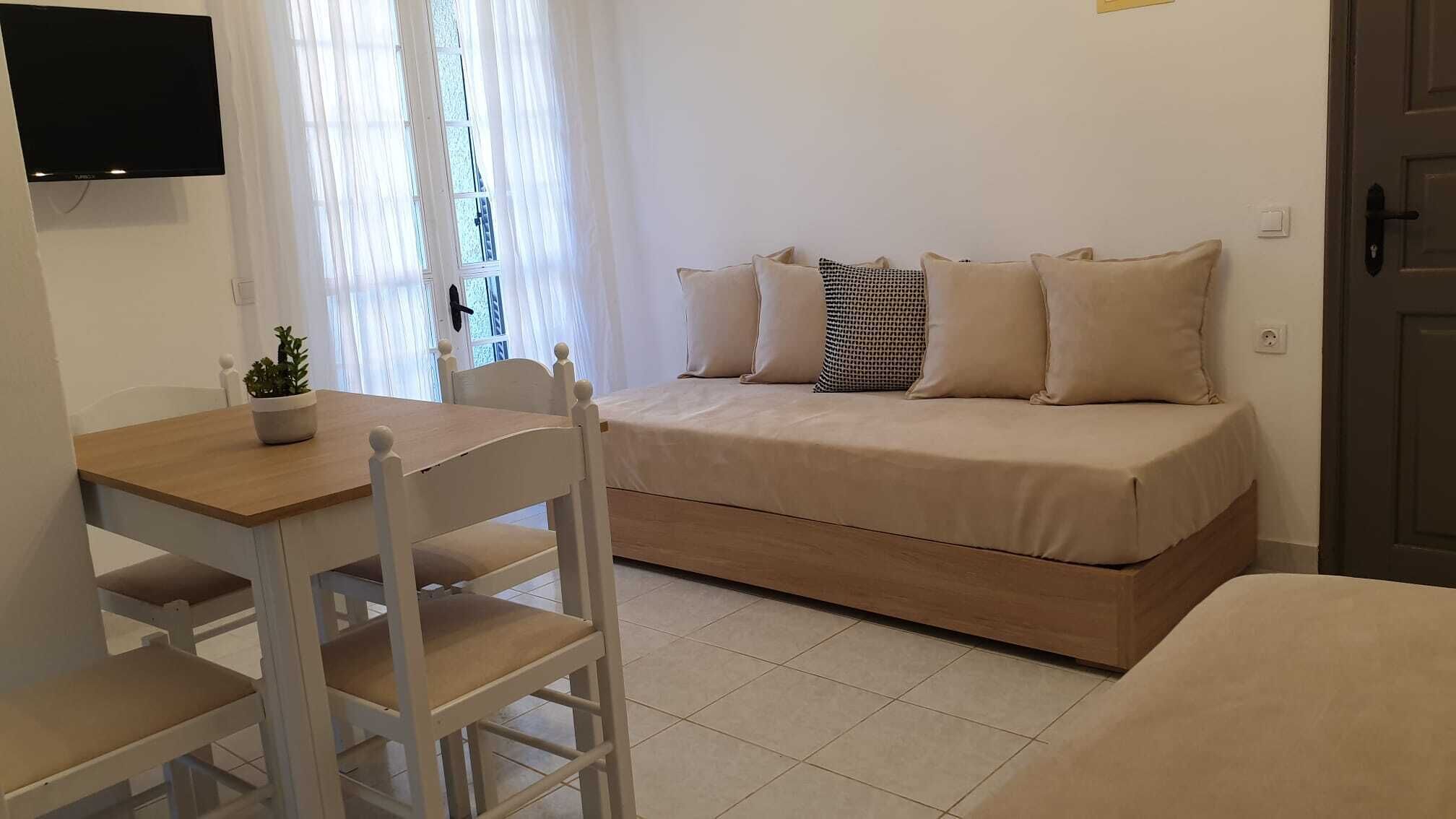A small, cozy room with a bed, a table, and a chair. There is a TV mounted on the wall and a window with curtains.