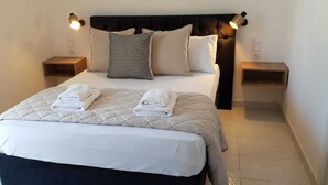 Een net opgemaakt bed met een gewatteerde dekbedovertrek, twee kussens en twee opgevouwen handdoeken. Er zijn twee nachtkastjes met lampen, en een donker hoofdbord.