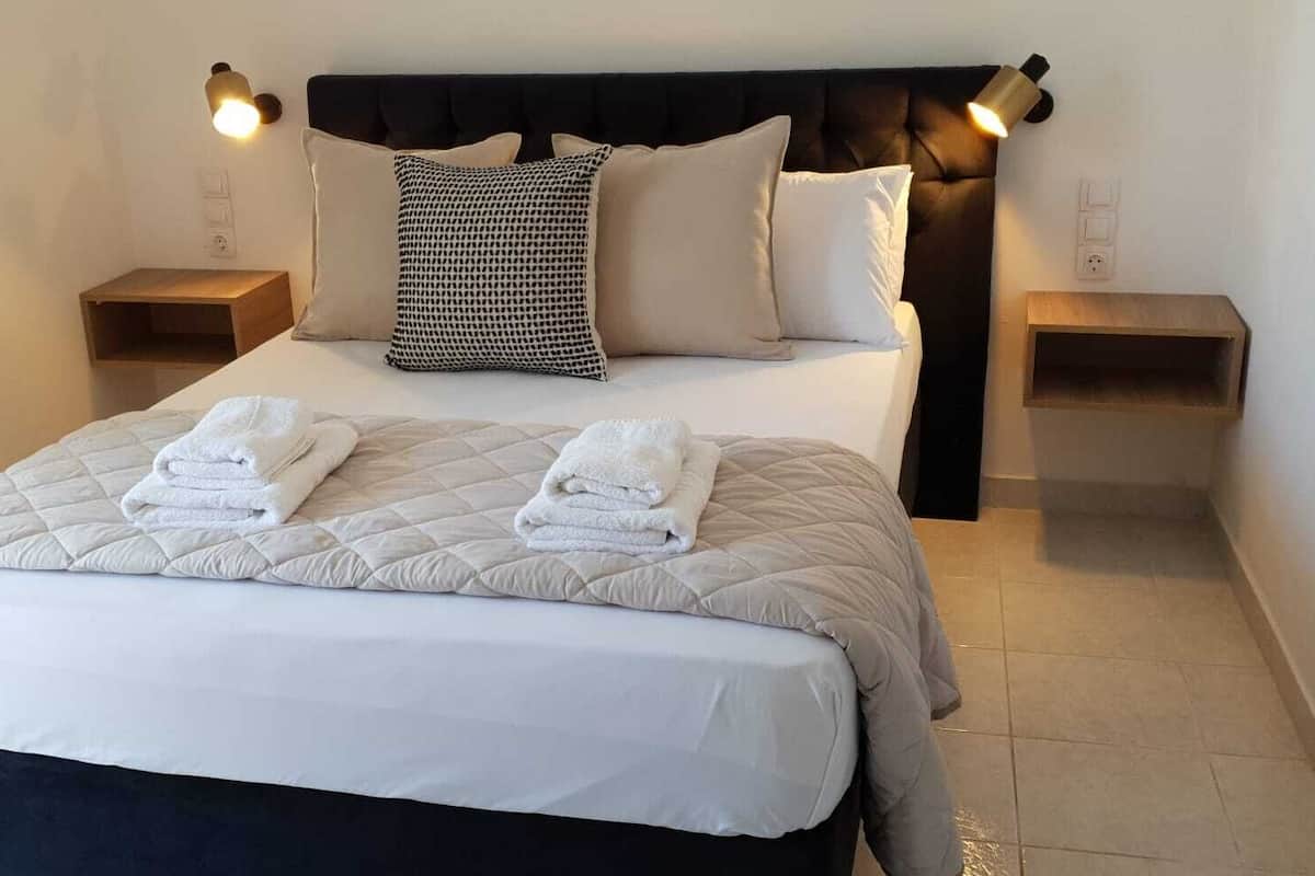 A neatly made bed with a quilted duvet, two pillows, and two folded towels. There are two bedside tables with lamps, and a dark headboard.