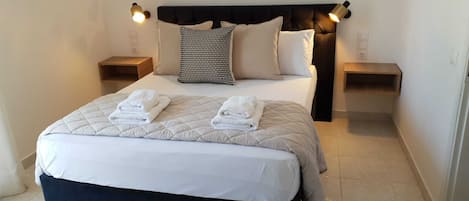 Una cama bien hecha con un edredón acolchado, dos almohadas y dos toallas dobladas. Hay dos mesitas de noche con lámparas, y un cabecero oscuro.