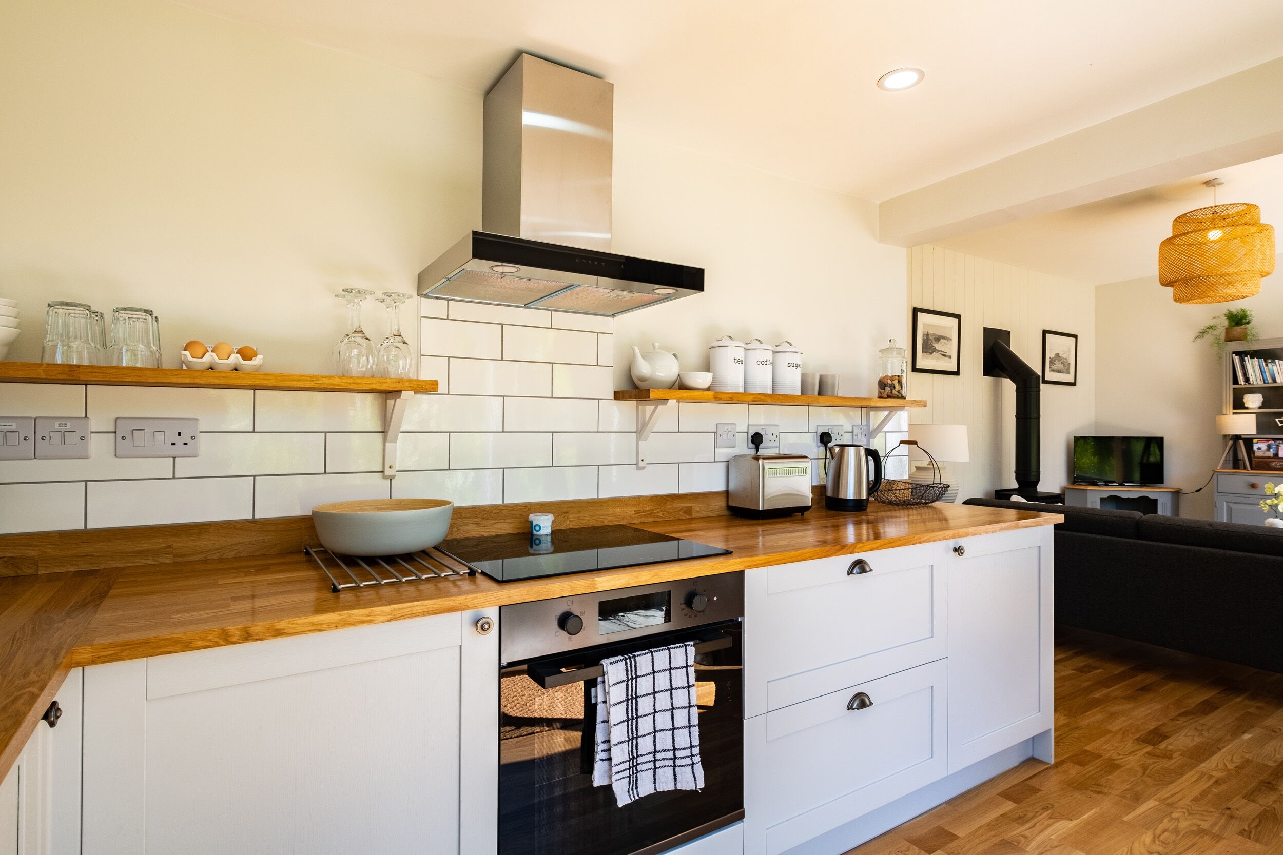 Een moderne keuken met witte kasten, een houten aanrecht en een ingebouwde oven. Op het aanrecht staan planken met glazen potten en een waterkoker.