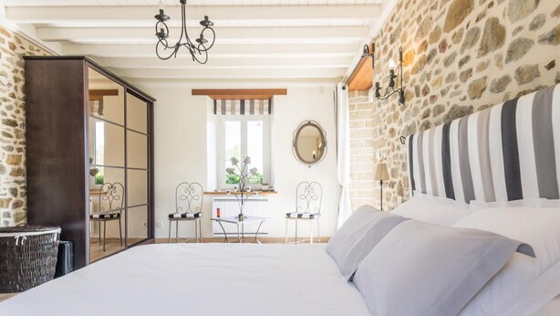 Un dormitorio con pared de piedra, una cama con ropa de cama blanca, una araña y vistas al exterior.
