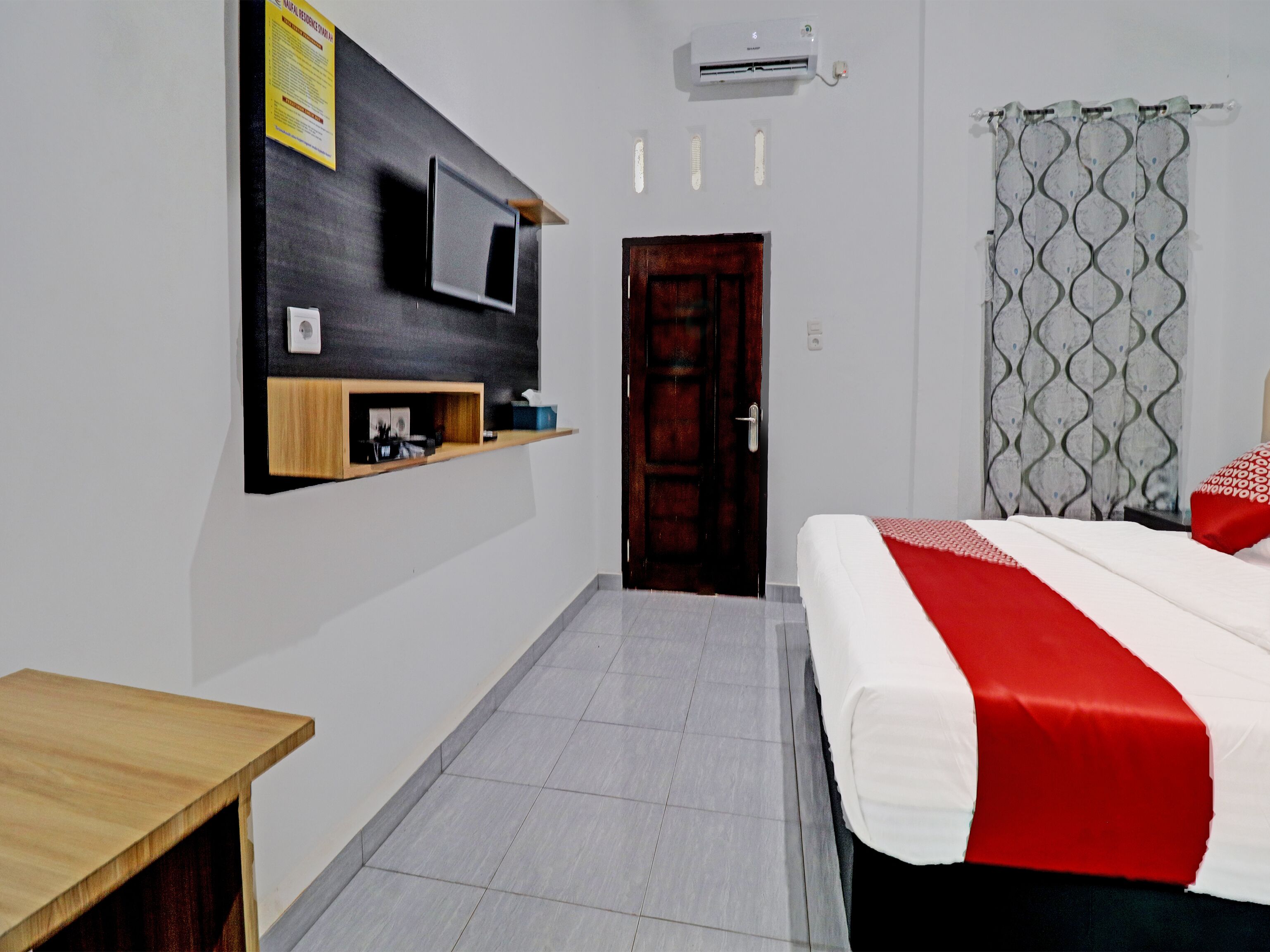 A hotel room with a bed, a TV mounted on the wall, a wooden shelf, a door, and a curtain.