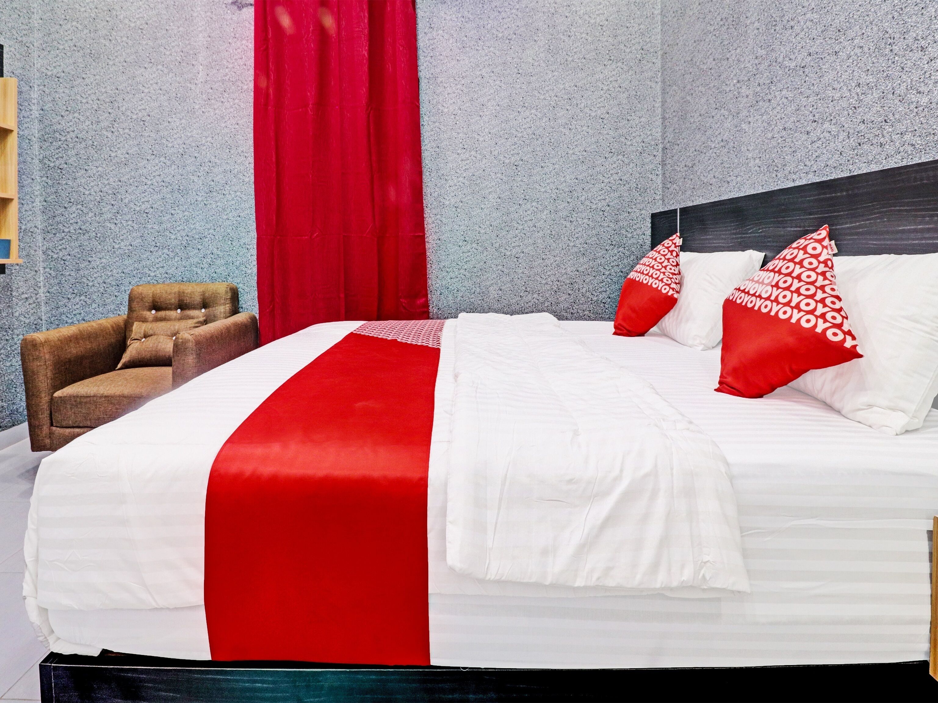 Una cama bien hecha con una colcha blanca y roja, un sillón marrón y una cortina roja.