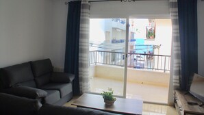 A living room with a sofa, a coffee table with a plant, and a sliding glass door leading to a balcony with a railing and a view of buildings.