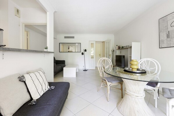 A modern living space with a glass-top dining table, white chairs, a sofa, and a flat-screen TV.