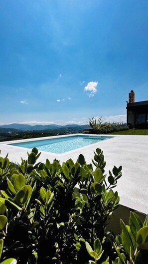Une piscine offrant une vue dégagée sur les montagnes lointaines, entourée de verdure et bordée d’un bâtiment moderne.