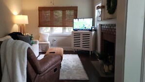 A living room with a brown leather sofa, a white armchair, a white fluffy rug, a television on a stand, a fireplace, and a window with blinds.