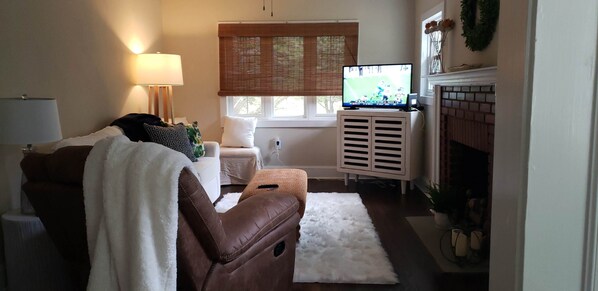 A living room with a brown leather sofa, a white armchair, a white fluffy rug, a television on a stand, a fireplace, and a window with blinds.
