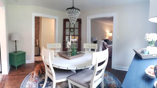 A dining area with a round table, white chairs, a chandelier, and a view of the outdoors through a window.