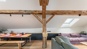 Quarto estilo loft com vigas de madeira aparentes, área de refeições com mesa e bancos de madeira, sofá e claraboia.