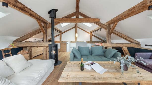 Uma sala de estar estilo loft com vigas de madeira aparentes, uma lareira central, um sofá, uma mesa de centro e um espaço para refeições.