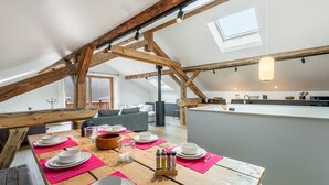 Uma área de jantar com mesa de madeira posta para quatro, cozinha ao fundo e teto inclinado com vigas aparentes.
