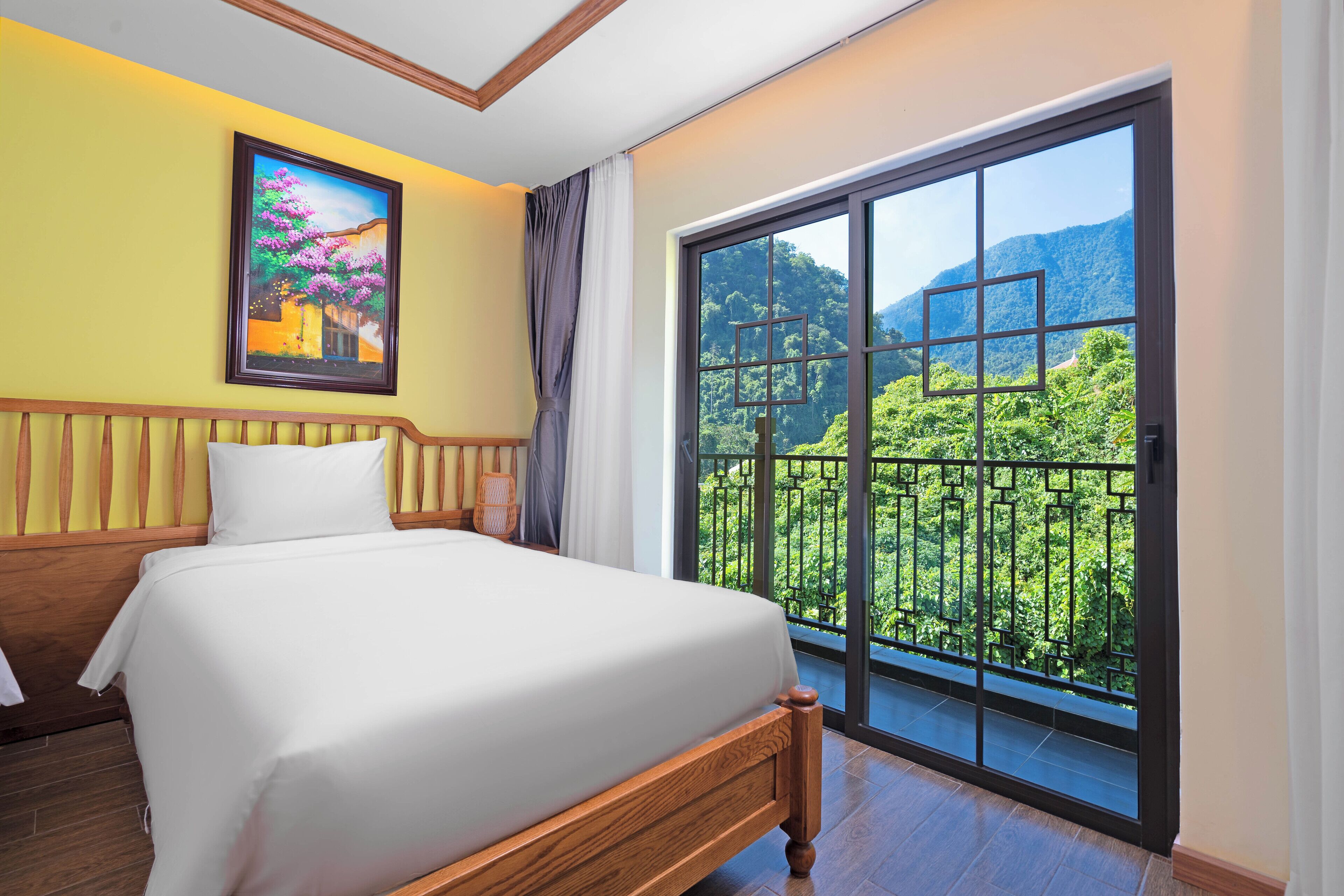 A hotel room with a bed, a painting on the wall, and a balcony with a view of greenery and mountains.