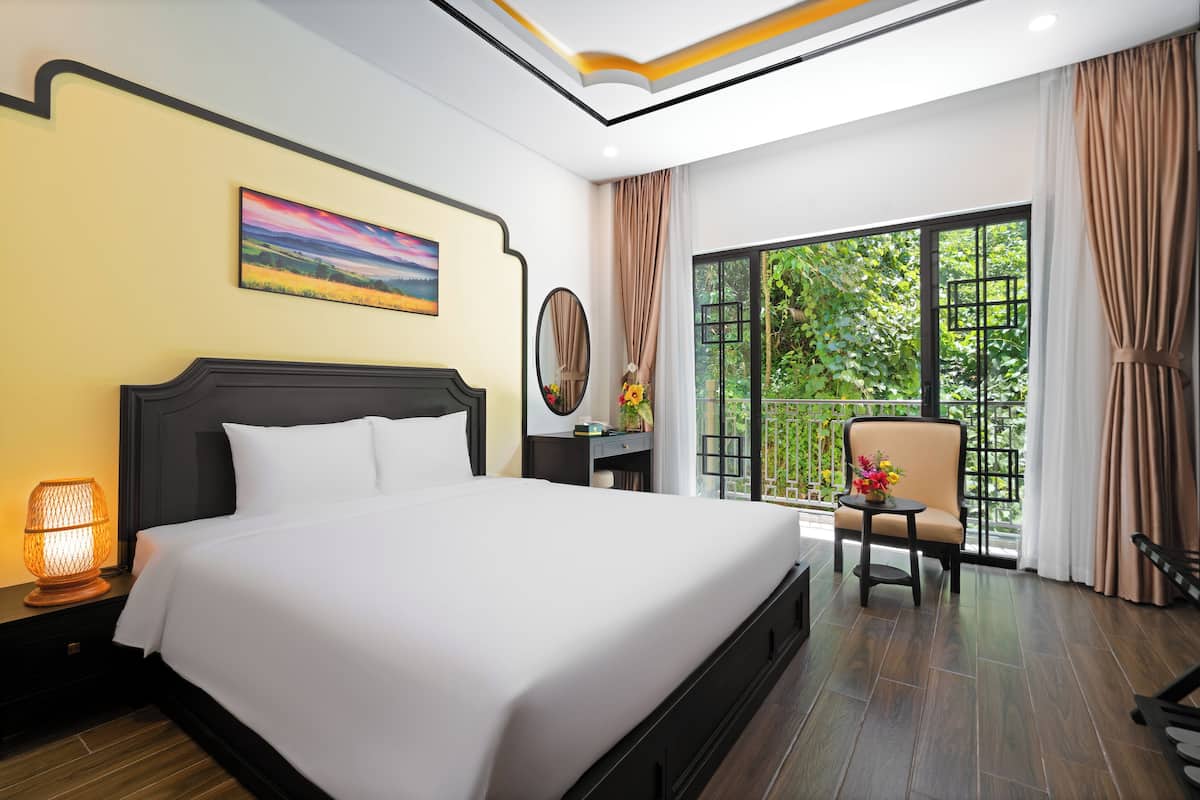 Une chambre d’hôtel avec un grand lit, une table de chevet avec une lampe, une chaise, une fenêtre donnant sur un paysage verdoyant et un tableau au mur.