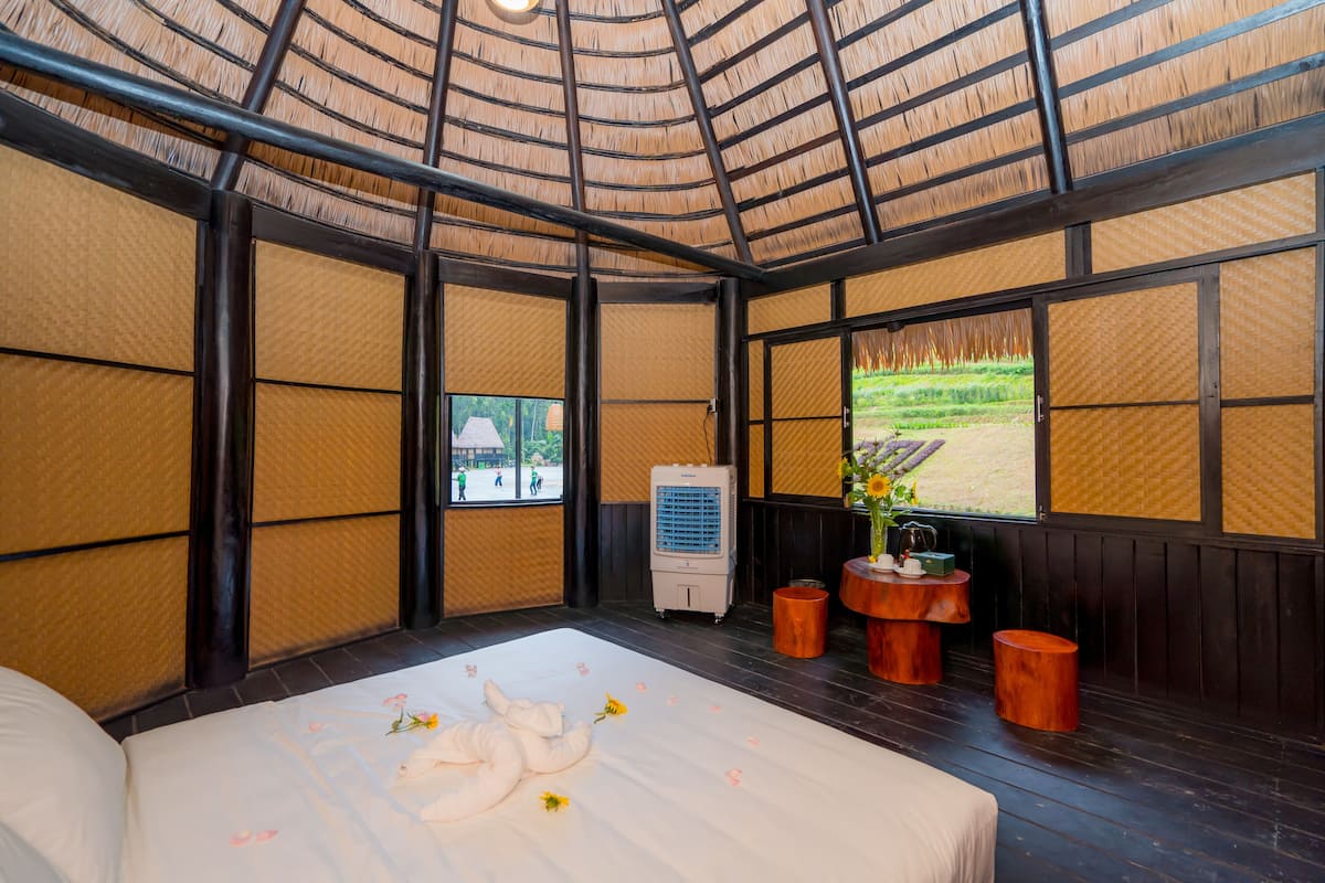 Une pièce de style japonais traditionnel, avec un sol recouvert de tatamis, un lit avec une couvre-lit blanche, une petite table ornée d’un vase et une fenêtre donnant sur un jardin.