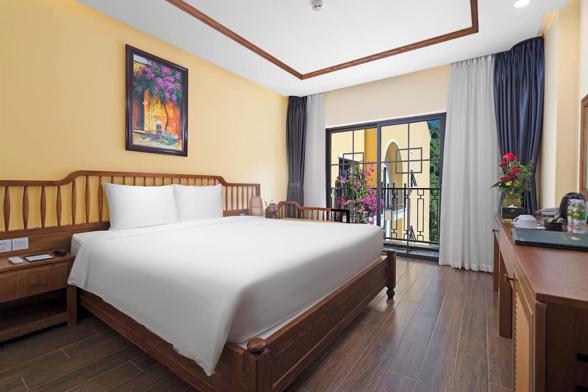 Une chambre d’hôtel avec un grand lit, une tête de lit en bois, une table de chevet, un bureau avec une chaise, un tableau au mur et un balcon avec vue.