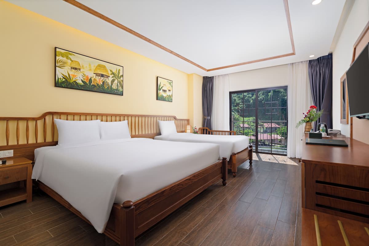 Une chambre d’hôtel avec deux lits, une tête de lit en bois, une table de chevet, un bureau avec une chaise, une télévision et un balcon donnant sur un espace verdoyant.