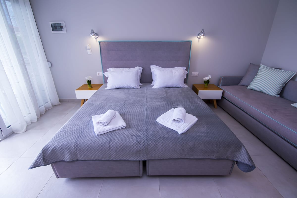 Ein Doppelbett mit grauer gesteppter Bettdecke und zwei weißen Kissen, flankiert von Nachttischen mit Lampen, ein graues Sofa mit einem Streifen-Kissen und ein Fenster mit durchscheinenden Vorhängen.