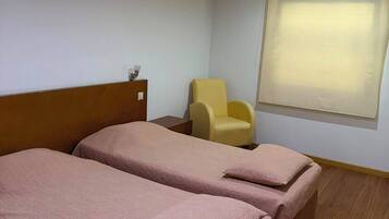 A hotel room with two beds, a wooden headboard, a small table, a chair, and a window with blinds.