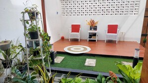 A patio area with a wooden floor, green artificial turf, white wicker chairs with red cushions, and a white lattice wall. - EXPRESS O 91277 Atiq Homestay Syariah (Serang)