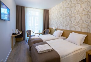 A hotel room with two beds, a desk with a chair, a TV, and a window with curtains. - Amber Sea Hotel (Jurmala)