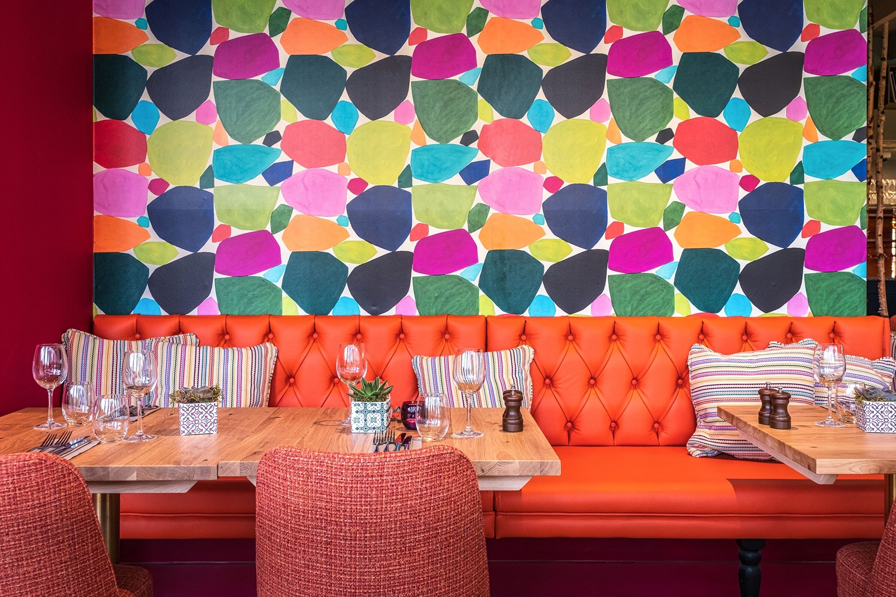 A vibrant restaurant interior with a colorful abstract wall, orange cushioned seating, and wooden tables set with wine glasses and cutlery.