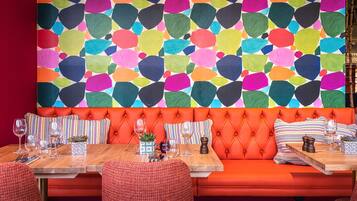 A vibrant restaurant interior with a colorful abstract wall, orange cushioned seating, and wooden tables set with wine glasses and cutlery.