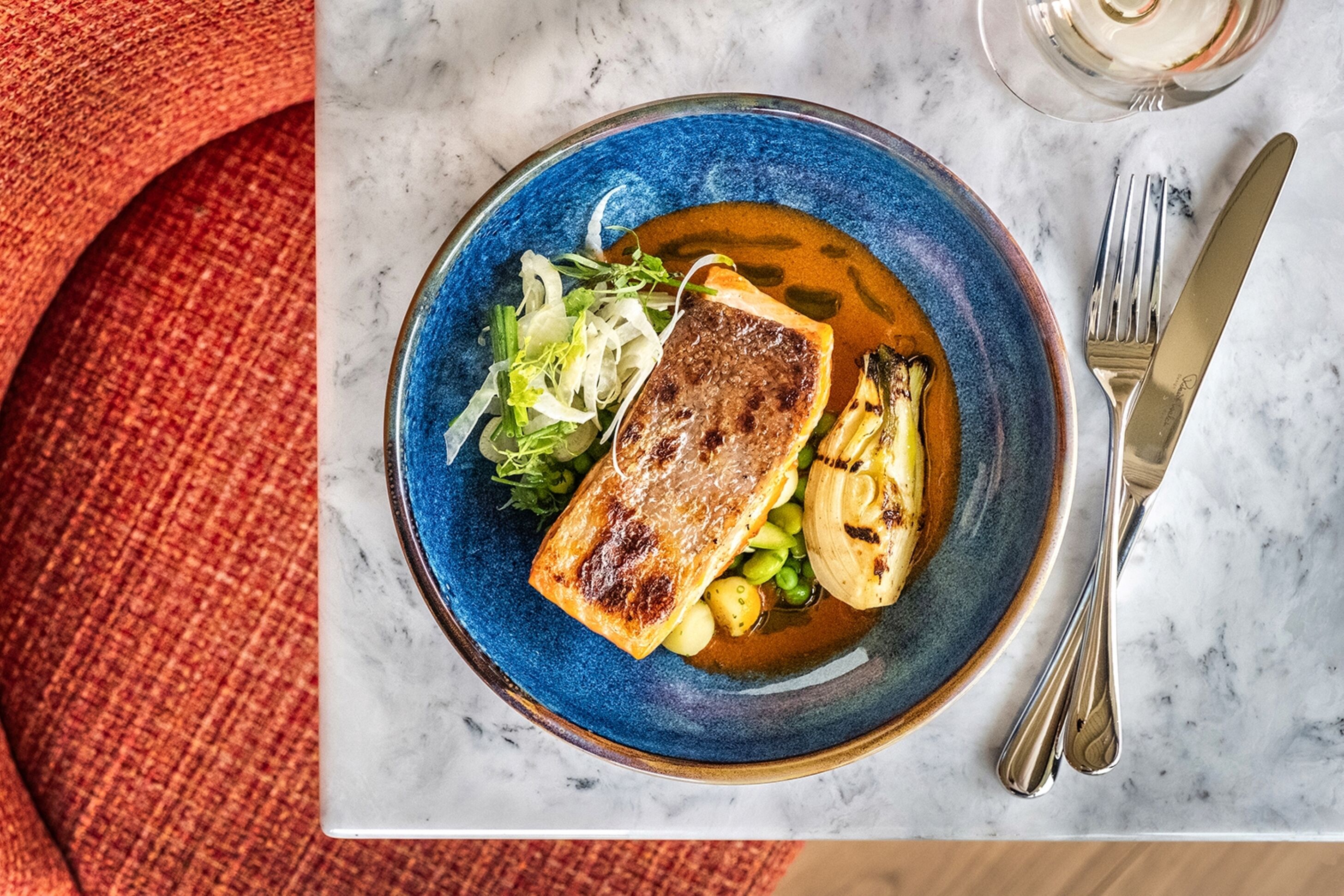 A plate of food with a piece of grilled meat, vegetables, and a blue bowl on a marble surface.