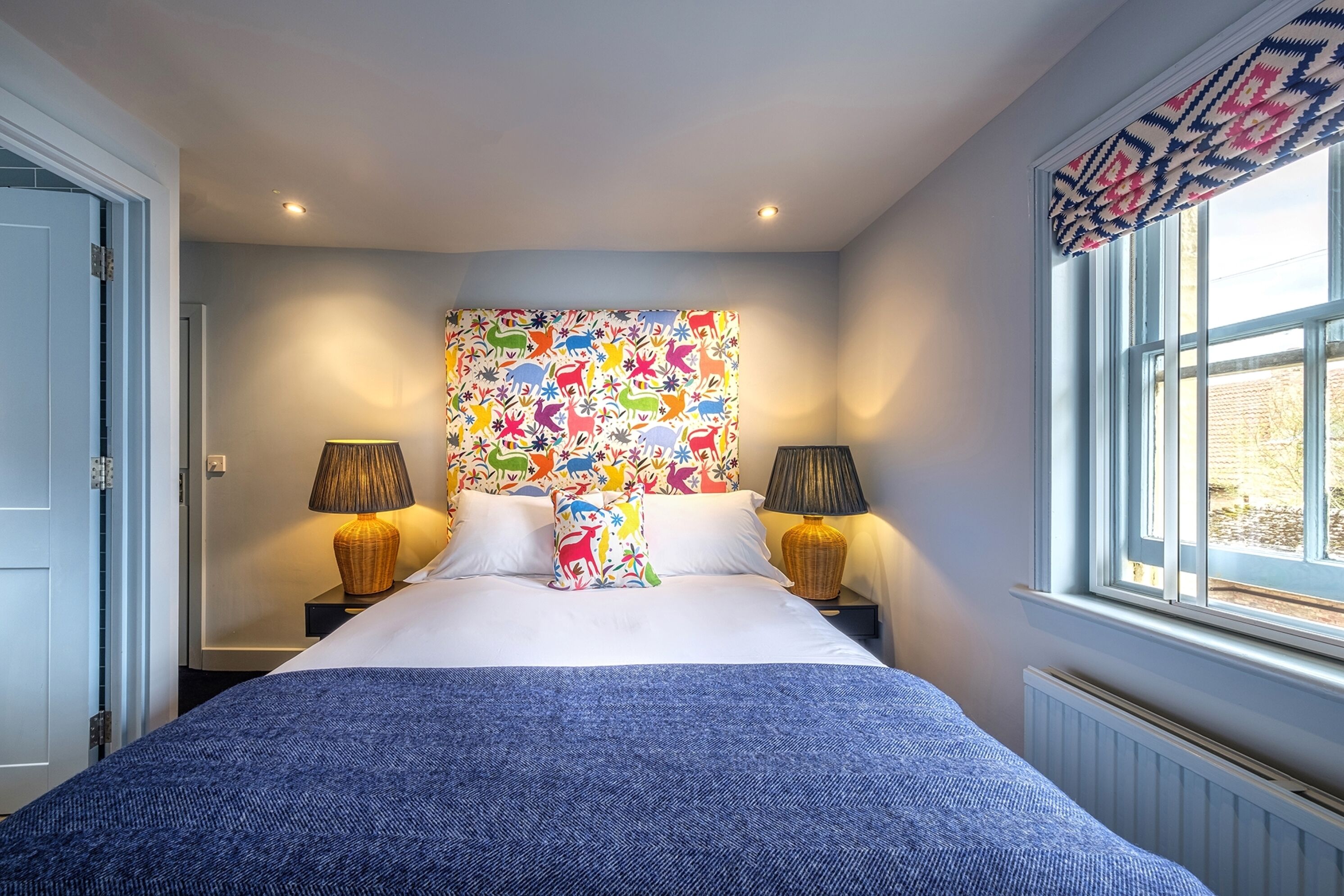 Un dormitorio con una cama grande, dos mesitas de noche con lámparas, un cuadro de pared colorido y una ventana con vistas.
