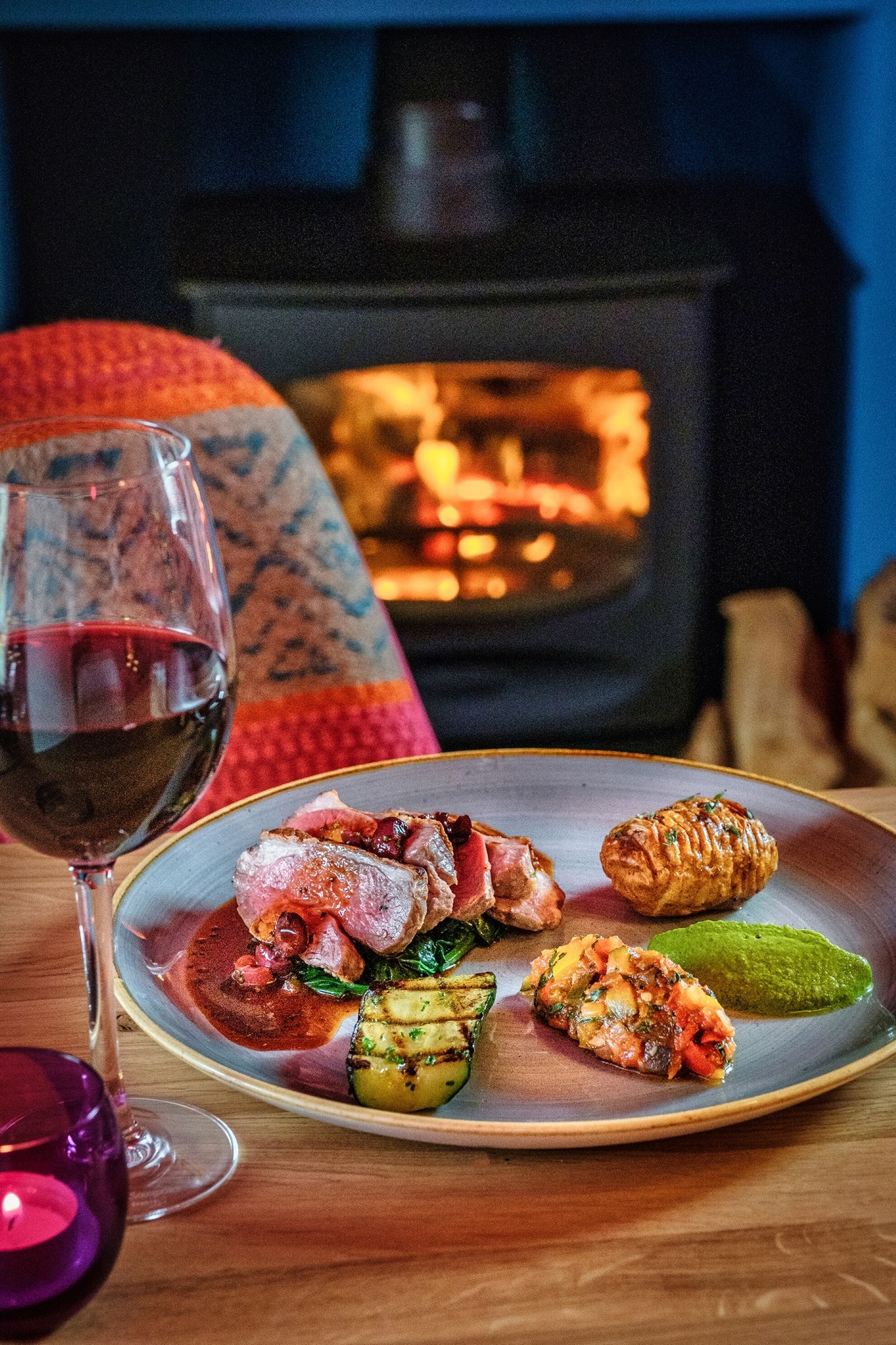 Une table à manger avec une assiette de nourriture, un verre de vin rouge et une cheminée chaleureuse en arrière-plan.