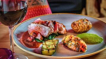Une table à manger avec une assiette de nourriture, un verre de vin rouge et une cheminée chaleureuse en arrière-plan.