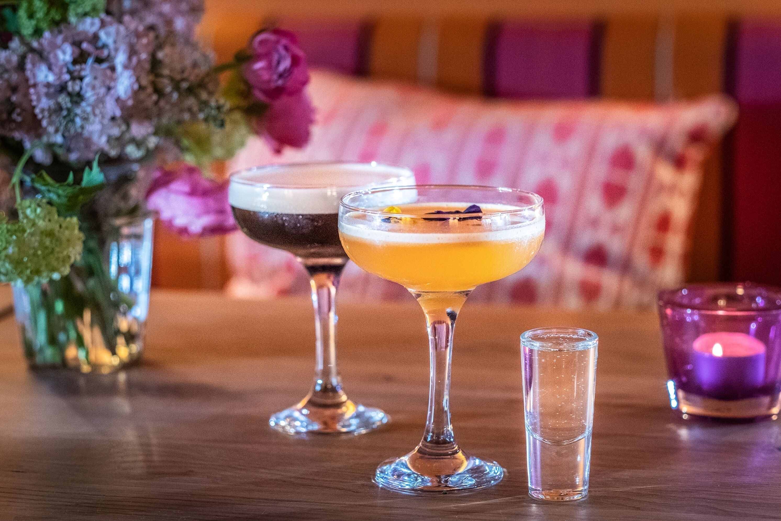 Three glasses of beverages on a wooden table, with a candle and flowers in the background.