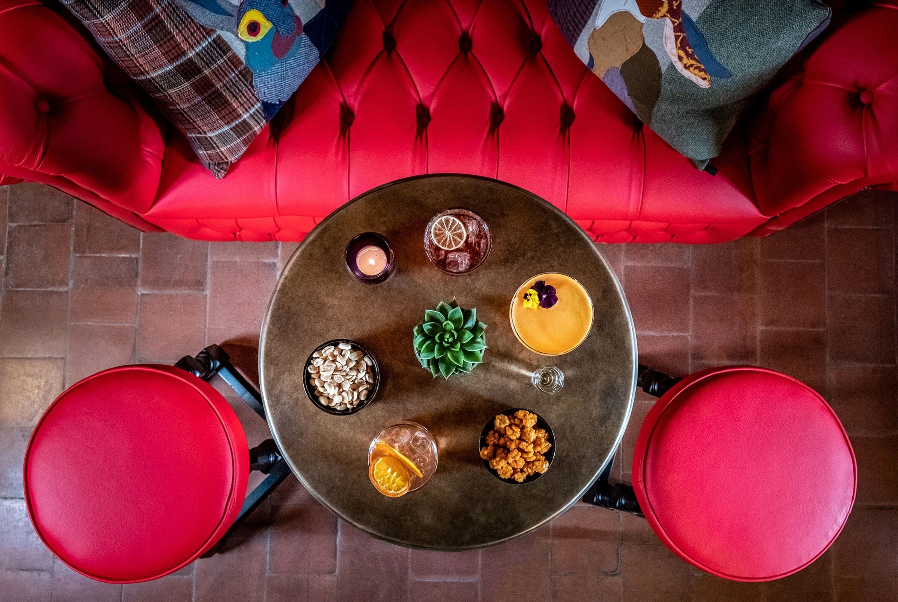 Una mesa redonda con comida y bebidas variadas, rodeada de sillas rojas, sobre un sofá rojo con cojines estampados.
