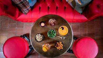 Una mesa redonda con comida y bebidas variadas, rodeada de sillas rojas, sobre un sofá rojo con cojines estampados.