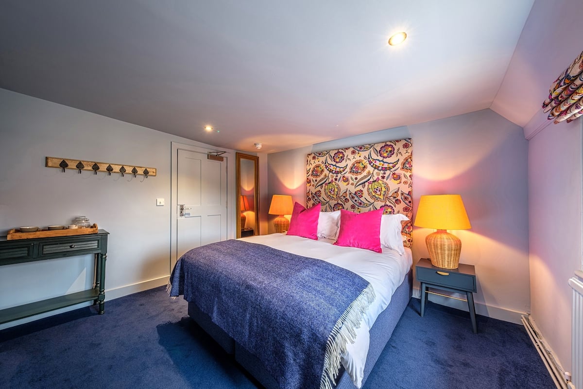 A bedroom with a bed, bedside lamps, a headboard with a floral pattern, a wooden coat rack, and a nightstand.