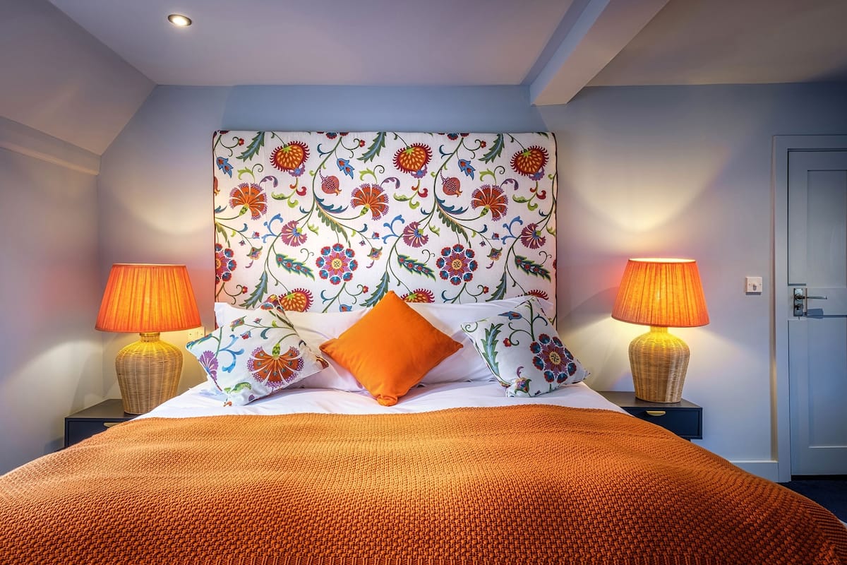 A neatly made bed with an orange blanket, two bedside lamps, and a floral-patterned headboard.