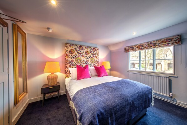 A bedroom with a large bed, two bedside lamps, a patterned headboard, a window with a view, and a radiator. - The Carpenters Arms (Cambridge)