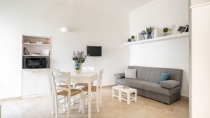 Ein modernes Wohnzimmer mit einem grauen Sofa, einem weißen Esstisch mit Stühlen, einem kleinen Fernseher und einem Regal mit Dekoration.