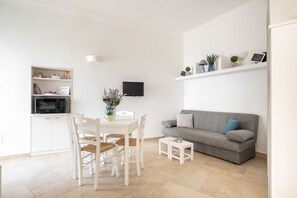 Un salon moderne avec un canapé gris, une table à manger blanche et des chaises, une petite télévision et une étagère ornée d’objets décoratifs.