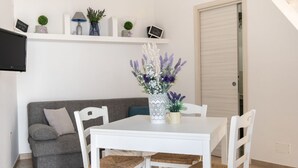 Ein weißer Esstisch mit vier Stühlen, ein graues Sofa, ein weißes an der Wand befestigtes Fernsehgerät und eine weiße Tür.