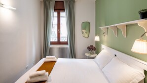 A hotel room with a bed, a window with curtains, a wall-mounted mirror, a small table with a vase, and a shelf with a plant. - GuestHost - Apartment located on the first floor of a building without a lift (15 steps at the entrance)In 37 sqm it accommodates 4 people, with a double bedroom and a sofa bed in the living room.Tram stop under the house at 150 meters.Parking on both wh (Padova)